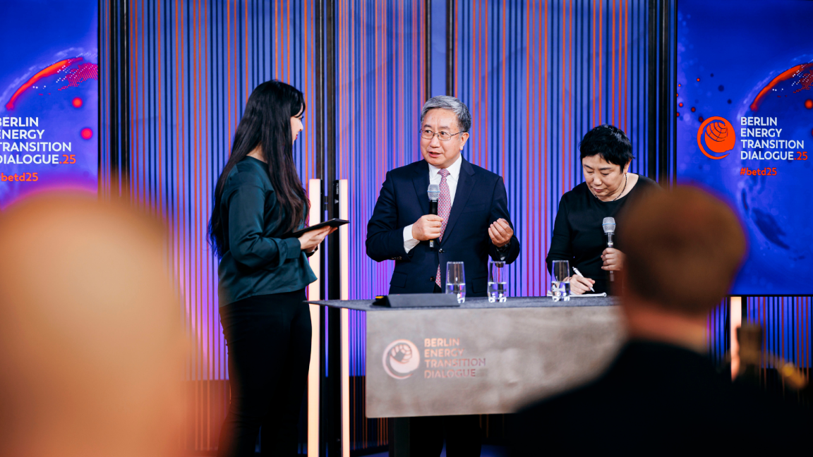 Three people talk to one another on a stage; behind them are two blue banners bearing the words ‘Berlin Transition Dialogue’.