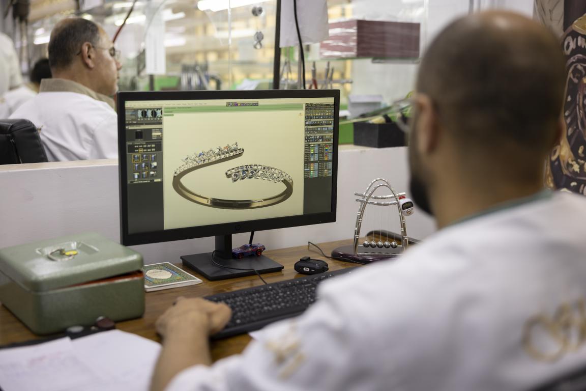 Bei Shawkat Alshami Jewellery arbeitet ein Schmuckdesigner, von hinten zu sehen, an seinem Schreibtisch und bedient die Tastatur. Auf dem Bildschirm ist das 3D-Modell eines offenen, spiralförmigen Goldrings zu sehen, der mit Reihen kleiner Edelsteine besetzt ist. Im Hintergrund arbeitet ein Kollege.