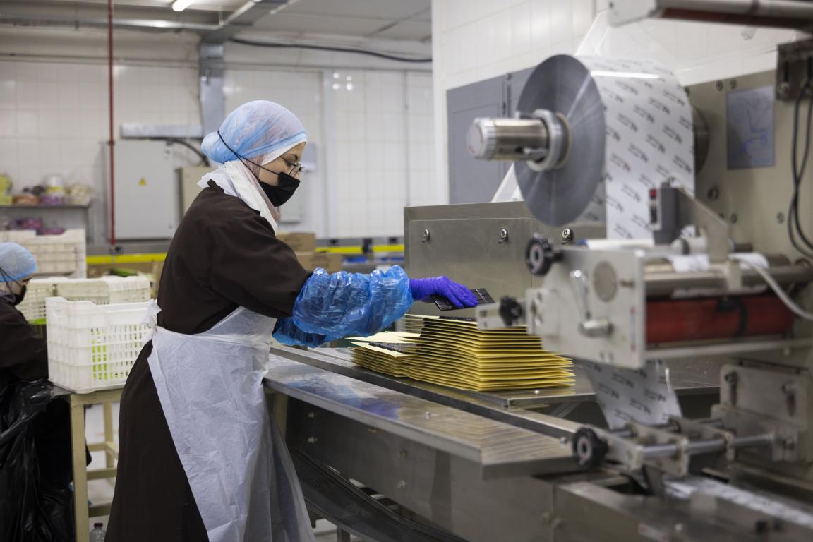 At the Tops factory, a factory worker in a blue hairnet, black face mask, brown smock, white apron, and purple gloves places a chocolate bar onto a stack of yellow separator sheets beside a stainless-steel wrapping machine loaded with a large roll of printed foil.