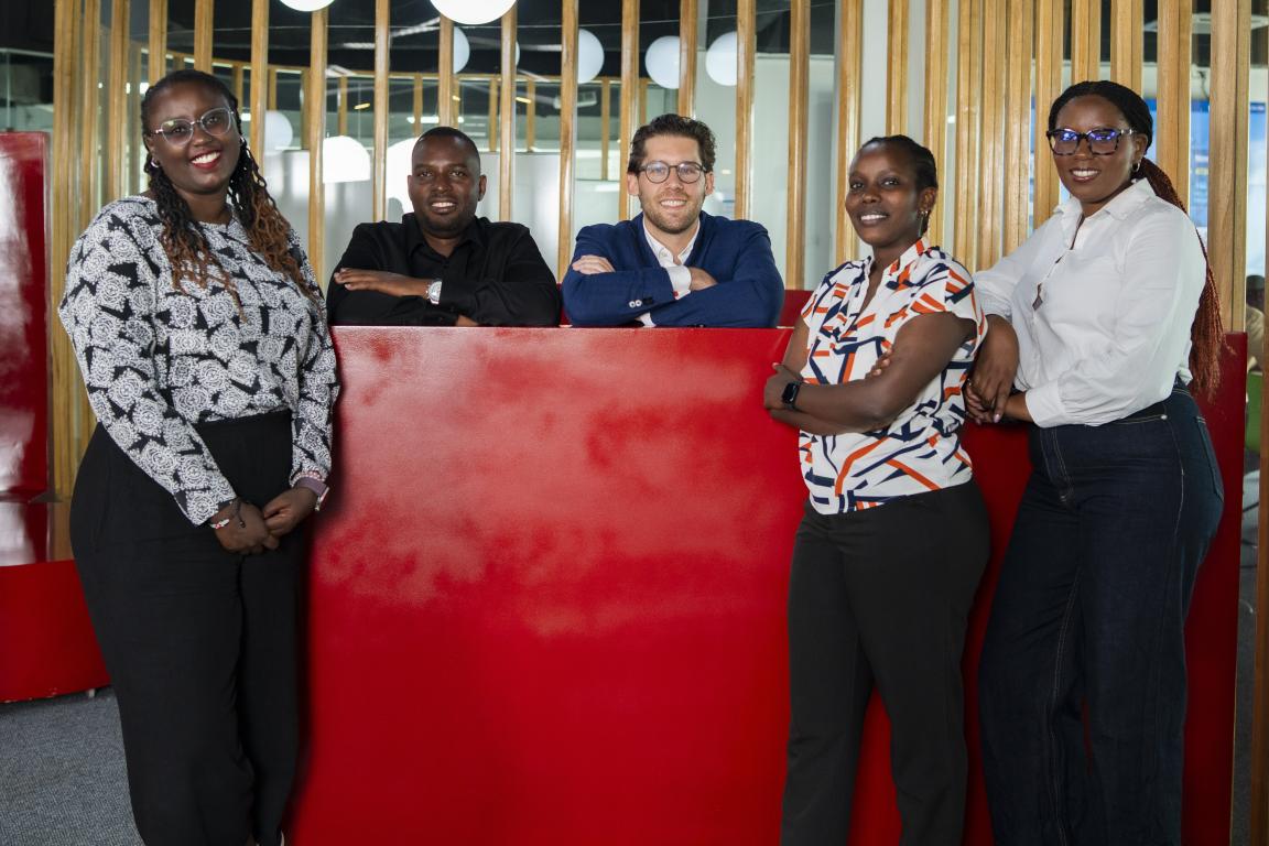 Five persons stand smiling behind a red divider in a modern office with wooden slats and round lights.