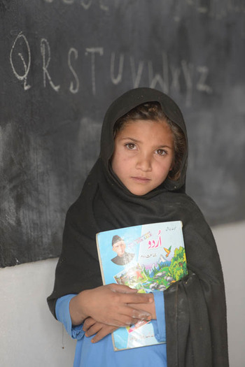 Ein Mädchen in einer Schuluniform steht vor einer Tafel und hält ein Schulbuch.