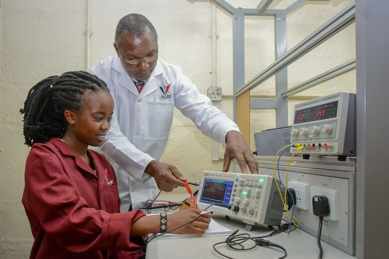 Ein Ausbilder erklärt einer jungen Frau ein technisches Gerät im Labor, während sie Notizen macht.© GIZ