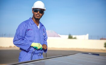 Ein Mann in Schutzkleidung hält ein Kabel in der Hand und steht hinter einem Solarpaneel.