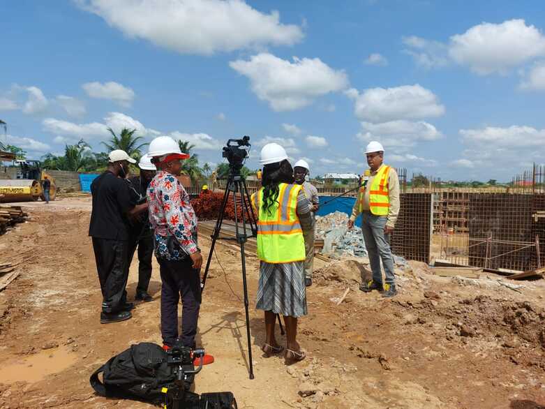 Ein Mann mit Helm und Weste gibt ein Kamerainterview. Er steht vor der Baustelle einer neuen Impfstoffproduktionsstätte.
