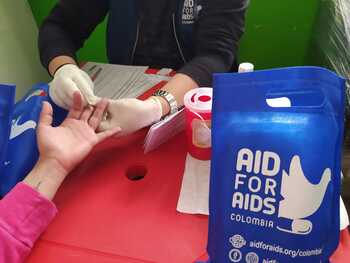 Einer Person wird eine Blutprobe am Finger entnommen. Auf dem Tisch steht eine Tüte von „Aid for Aids Colombia“.