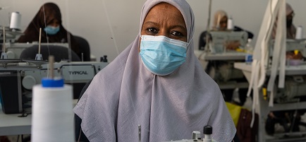 Eine Frau mit Gesichtsmaske sitzt an einer Nähmaschine und blickt in die Kamera.