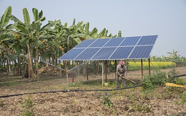 Ein Mann jätet Unkraut auf einer Wiese in Indien. Ein großes Solarpanel steht direkt hinter ihm, das eine solarbetriebene Bewässerungspumpe im Vordergrund antreibt. © SwitchON