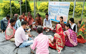 Eine Gruppe von Landwirt*innen sitzt in traditioneller indischer Kleidung auf einer Decke im Kreis auf dem Boden und hört einem Ausbilder zu, der in der Mitte der Gruppe sitzt. © SwitchON