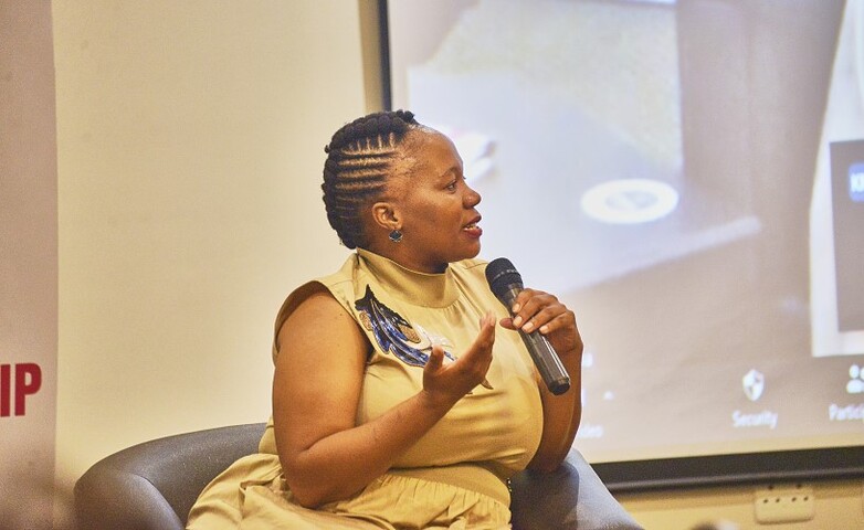 Eine Frau sitzt auf einem Stuhl, hält ein Mikrofon in der Hand und setzt zum Sprechen an.