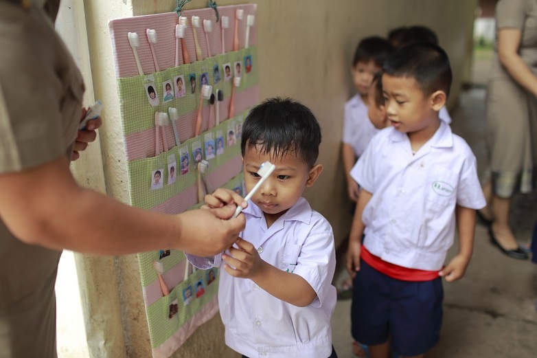 Ein Erwachsener gibt einem Kind eine Zahnbürste. Im Hintergrund befinden sich viele weitere Zahnbürsten in Taschen, auf denen Fotos von Kindern angebracht sind. Standort: Kambodscha. Copyright: GIZ.