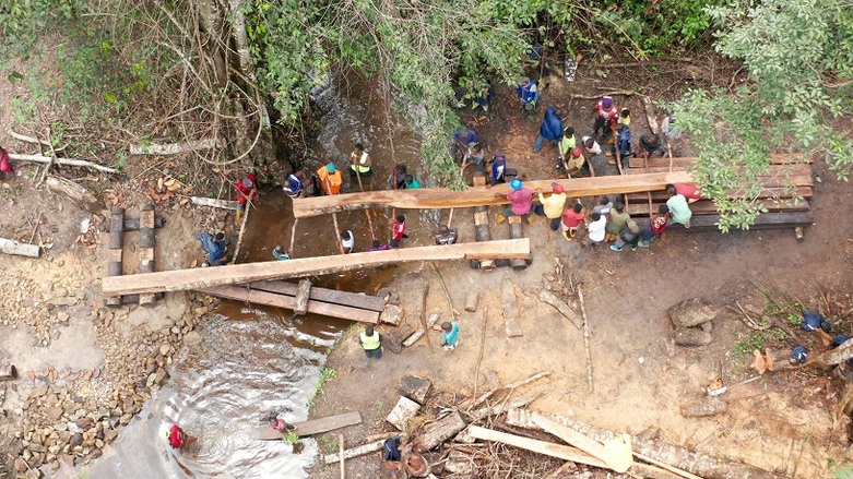Etwas dreißig Personen tragen große Holzbalken über einen Fluss. Copyright: John Healey
