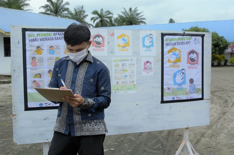 Eine Person füllt ein Dokument auf einem Klemmbrett aus. Im Hintergrund sind Plakate über Hygienevorschriften zu sehen. Standort: Indonesien. Copyright: GIZ.
