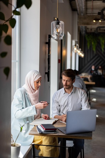 A woman with a hijab and a man are discussing in a café. In front of them is a laptop and stacks of notebooks. Copyright: GIZ