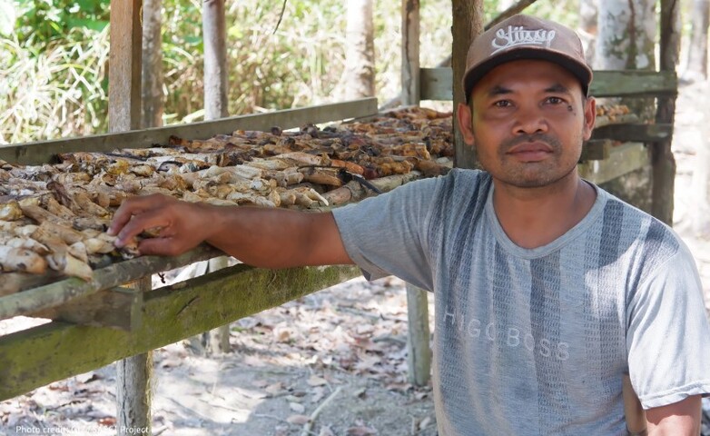 Ein Mann steht neben landwirtschaftlichen Erzeugnissen, die auf einem Holzgerüst trocknen. 