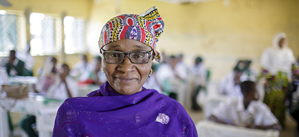 Eine Frau mit einem bunten Kopftuch und Brille steht in einem Klassenzimmer.