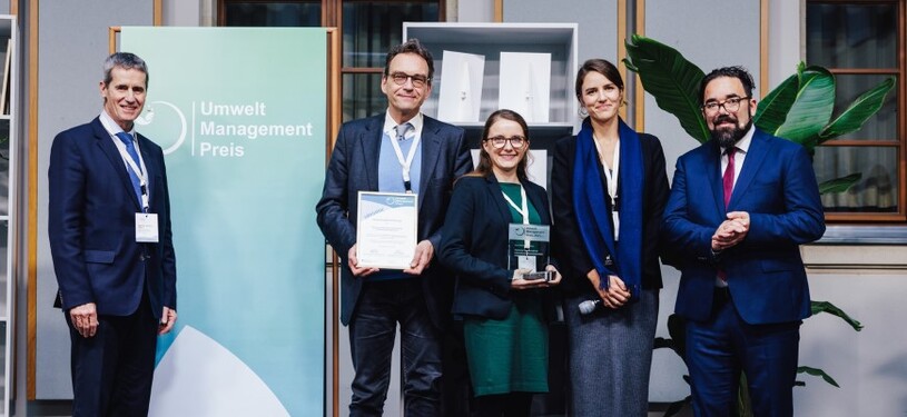 Eine Gruppe von Menschen in Geschäftskleidung steht vor einem grün-weißen Banner mit der Aufschrift ‘Umwelt Management Preis’ und hält eine Glas-Trophäe mit grünem Sockel.