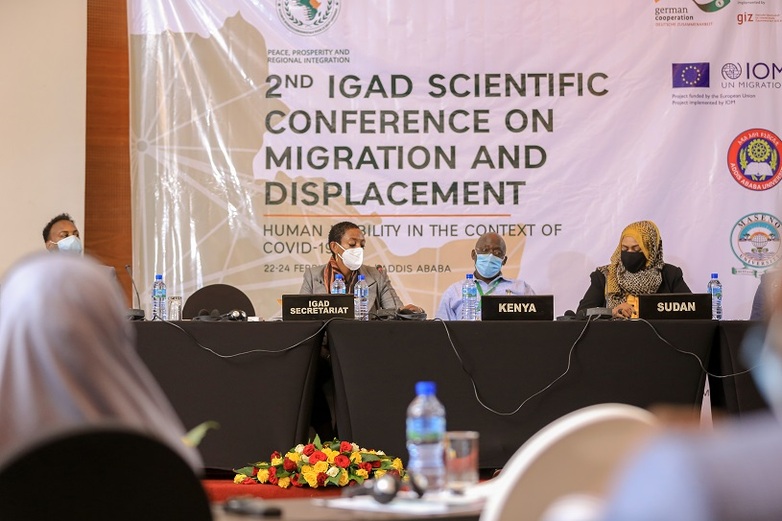 Personen mit Masken sitzen auf einer Bühne. Vor ihnen stehe n Schilder mit "Kenya" und "Sudan" etc. Hinter ihnen hängt ein Banner mit der Aufschrift "Second IGAD scientific conference on migration and displacement". Copyright: GIZ
