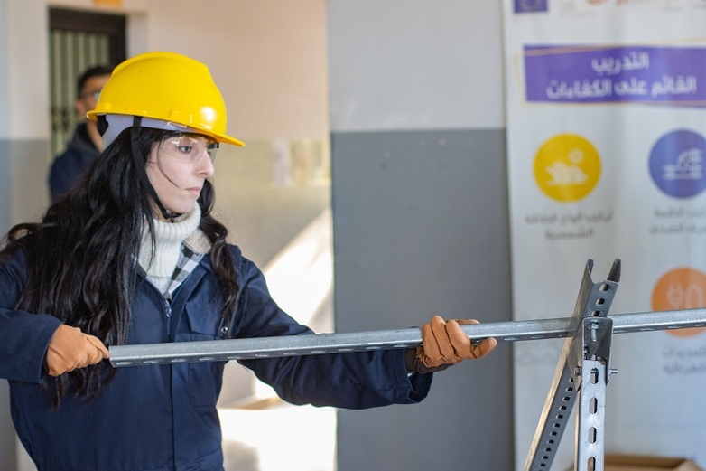 Eine junge Frau in Arbeitsschutzkleidung hält eine Stahlkonstruktion in der Hand.