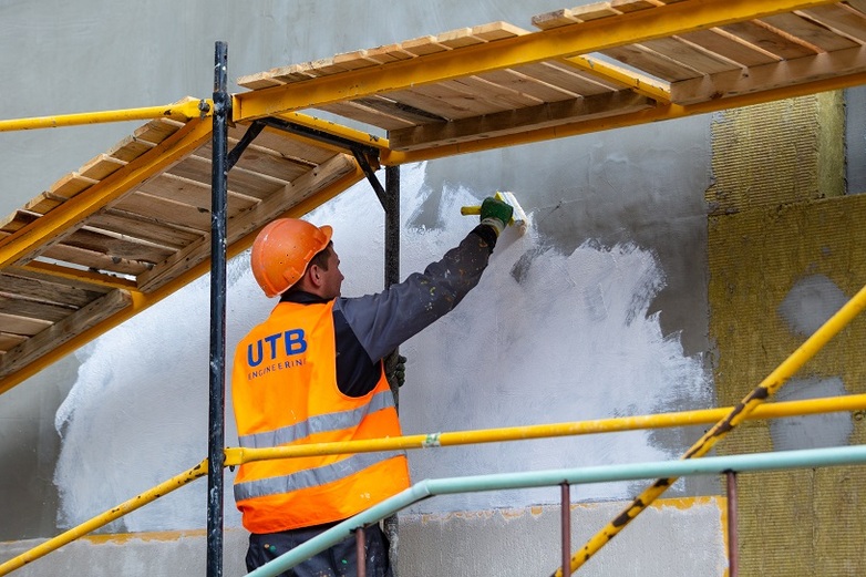 Eine Person in Warnweste und mit Schutzhelm streicht eine Wand mit weißer Farbe im Rahmen eines Renovierungsprozesses.