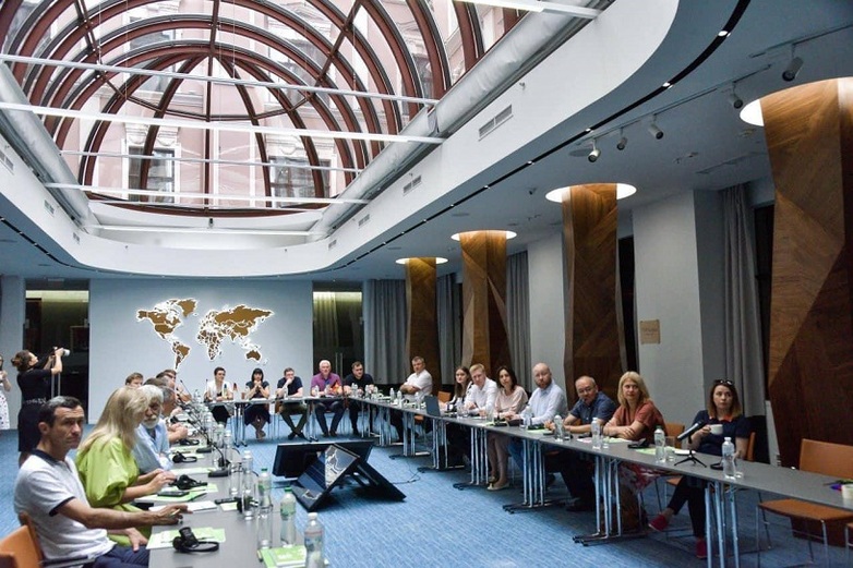 Viele Personen sitzen an einem langen, U-förmigen Tisch und besprechen sich.