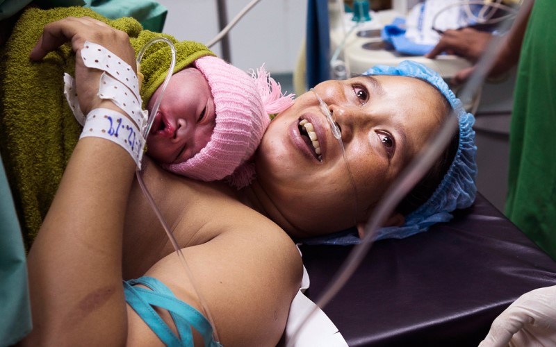 A young woman receiving medical care holds a newborn baby on her upper body.