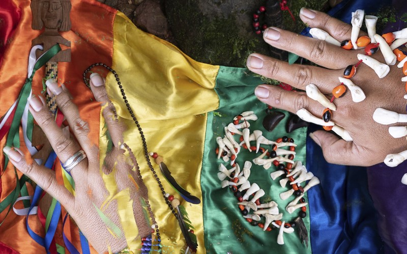 Zwei Hände und indigener südamerikanischer Schmuck auf einer Regenbogenfahne. 