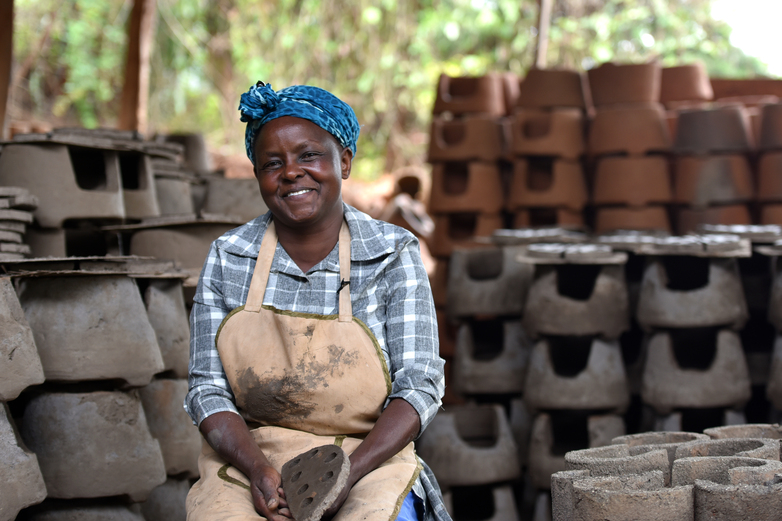 A woman wearing an apron is smiling as she sits next to the components required for producing stoves. Copyright: GIZ