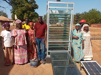 A group of men and women gather around a solar dryer in a village.