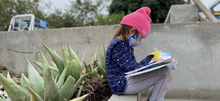 A girl with a face mask is sitting on a wall and writing in a notebook.