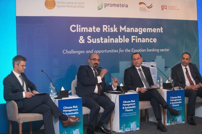 Four men in suits sit on a stage and discuss the topic of climate risk management and sustainable finance.
