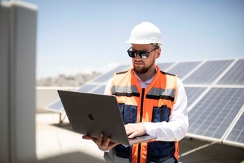 A man with sunglasses and a hard hat holds a laptop and stands in front of solar panels.
