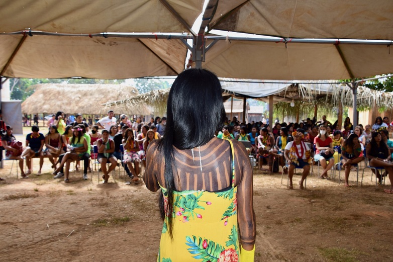 Vista das costas de uma mulher em pé na frente de mulheres indígenas em um evento. 