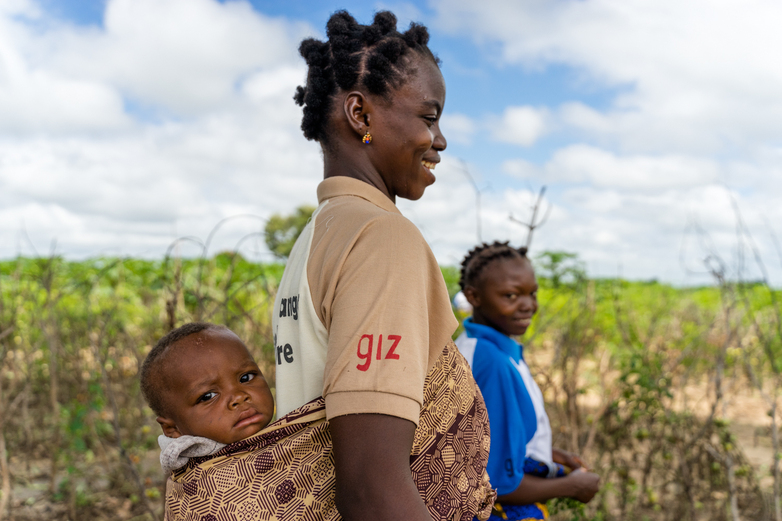 A woman stands before a field with a small child on her back, who is looking into the camera.