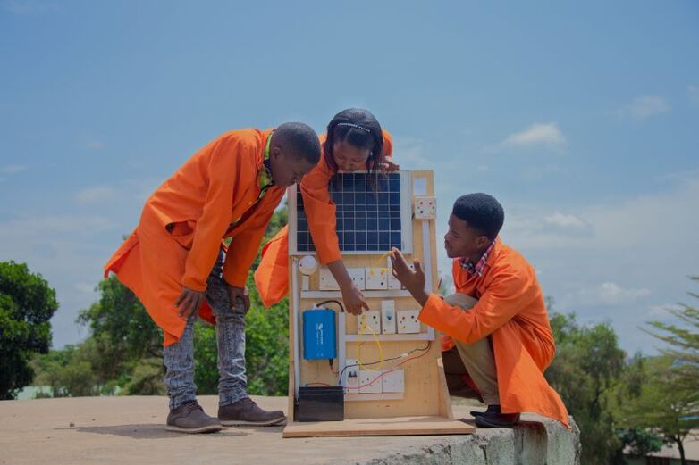 Three people in orange coats look at an electrical system connected to solar cells.
