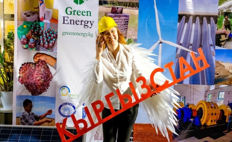 A young woman stands in front of a poster for a trade fair on green energy and holds an object resembling a lettering in her hand.