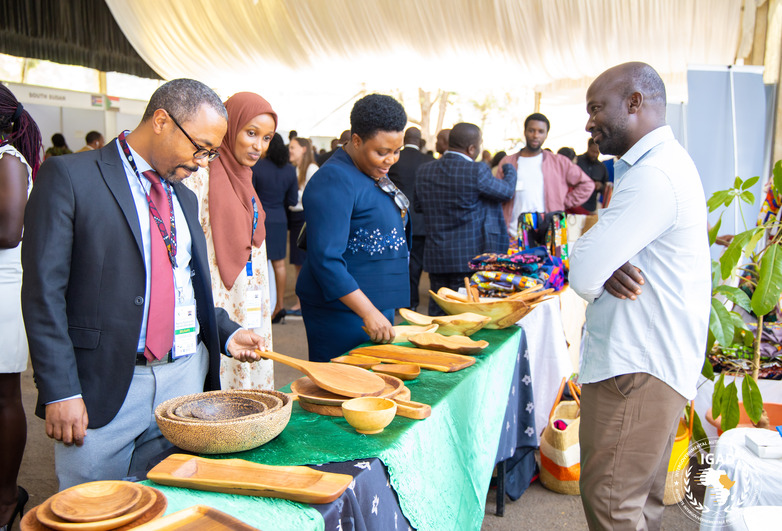 Participants in the Kampala Declaration meeting stand in front of a stand containing artisanal products, behind which a displaced entrepreneur stands. Copyright: IGAD 
