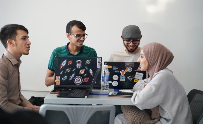 Four young individuals are sitting at a table with laptops and chatting.