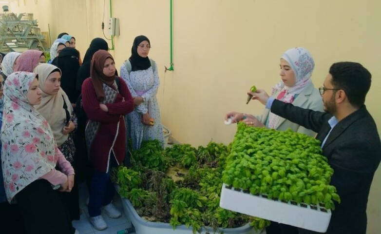 A man and a woman standing behind a large number of seedlings instruct a group of women on how to use them.