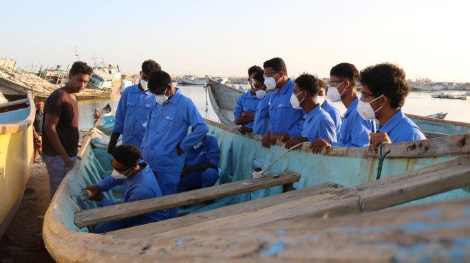 Young men in blue uniforms are trained to repair a boat.
