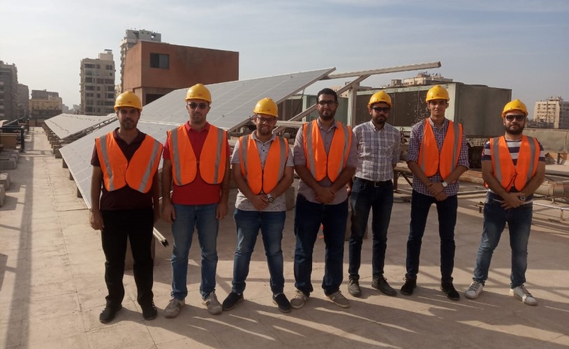 A group of men in protective clothing stand in front of a solar panel.