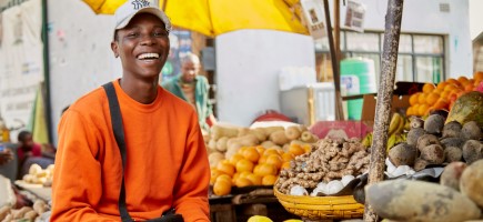 A man in a yellow sweater in front of a market stall with vegetables.