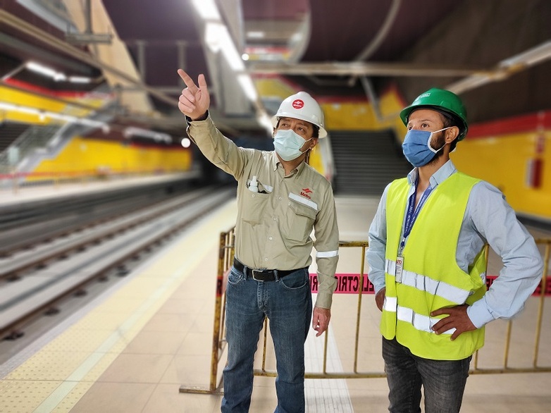 Two men wearing protective helmets and masks are standing on the platform of an underground train station. One of the men is pointing to the upper left-hand corner. Copyright: GIZ