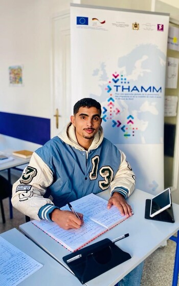 A young man attends a language class and sits at a desk while writing in a notebook. Behind him is a THAMM display stand.