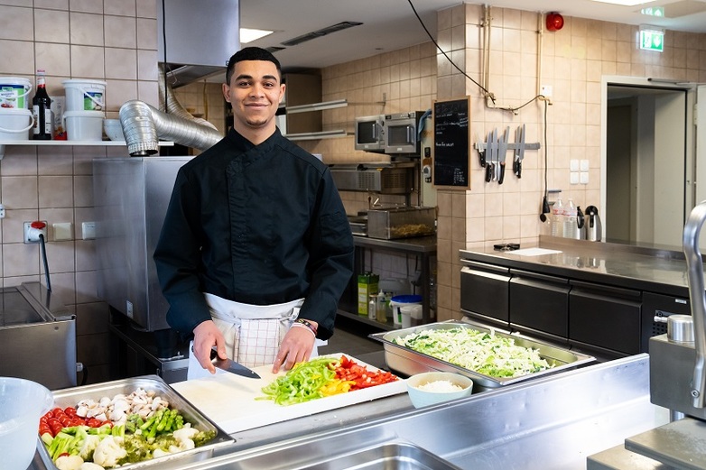 A young trainee stands smiling in a kitchen, chopping vegetables.