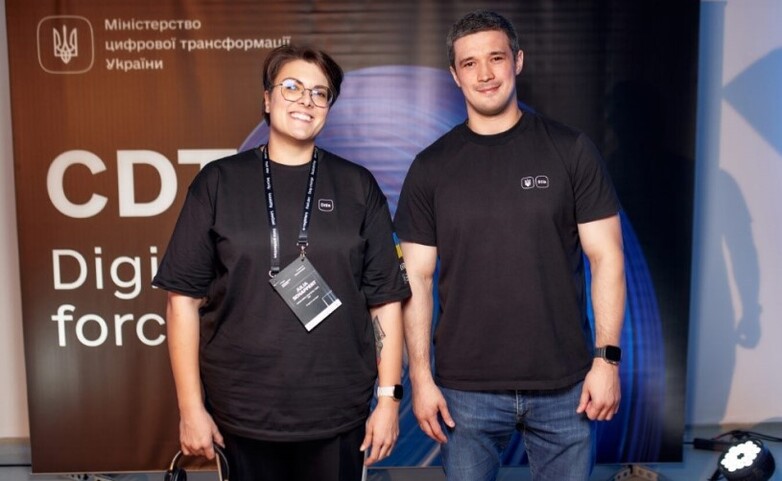 Two people stand in front of a banner of the Ministry of Digitalisation