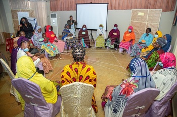 A crowd of people on a market square. Some carry large bowls and goods. Internally displaced women sit in a circle at a training course on basic entrepreneurial skills. A rope is stretched out between them.