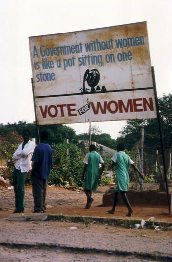 Four people under a sign that reads "A government without women is like a pot sitting on one stone - vote for women." Copyright: GIZ, Stefan Erber