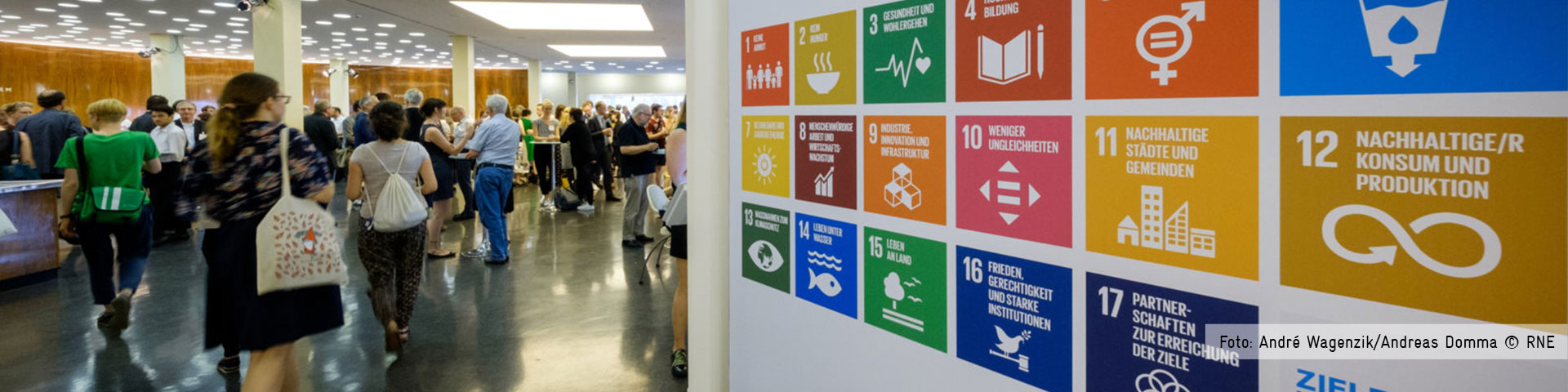 People at a conference walking and standing at the entrance of a building. On one side there is a wall displaying the 17 Sustainable Development Goals. Photo: André Wagenzik/Andreas Domma © RNE