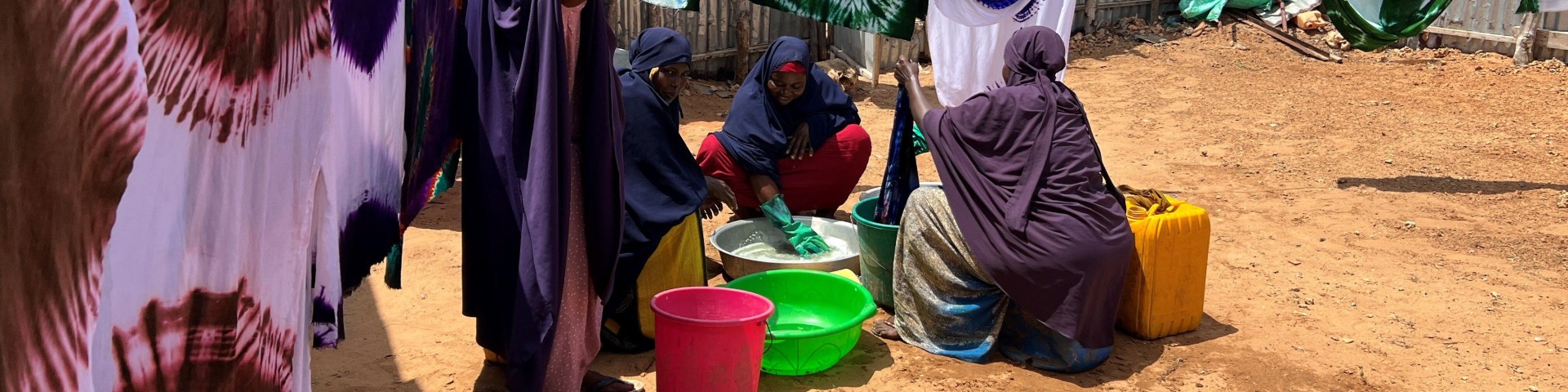 Four women tie-dye cotton fabrics. In the background, dyed fabrics are hung on a line to dry. The dyed fabrics are then sold on the market and later made into traditional Somali dresses.