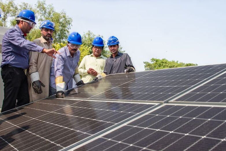 Five men in helmets and protective goggles in discussion alongside solar panels. 